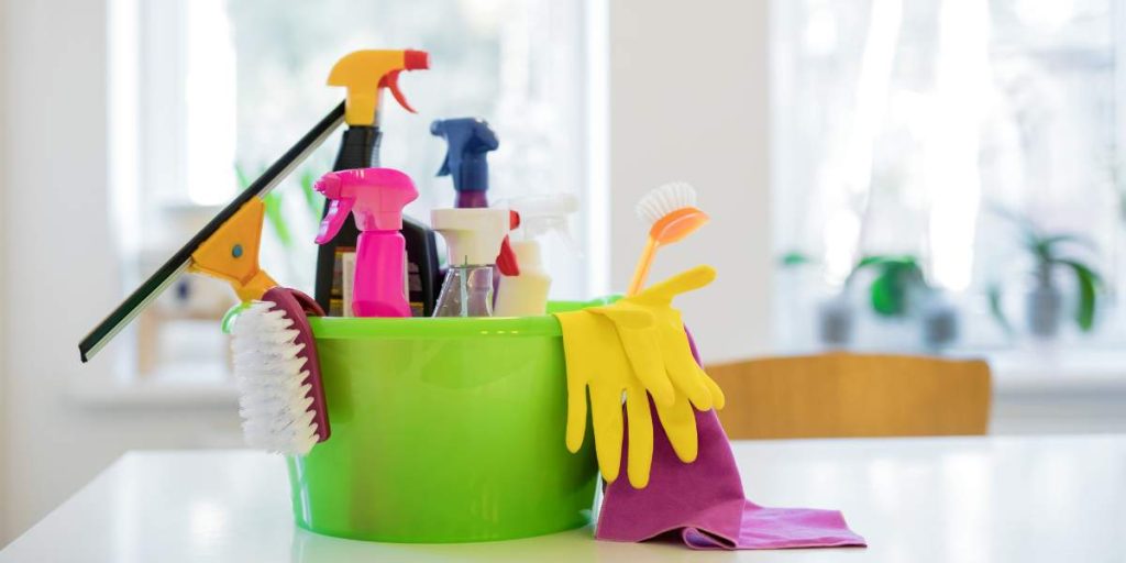 Rubber gloves, spray bottles, a squeegee, and other professional house cleaning supplies in a green bucket.